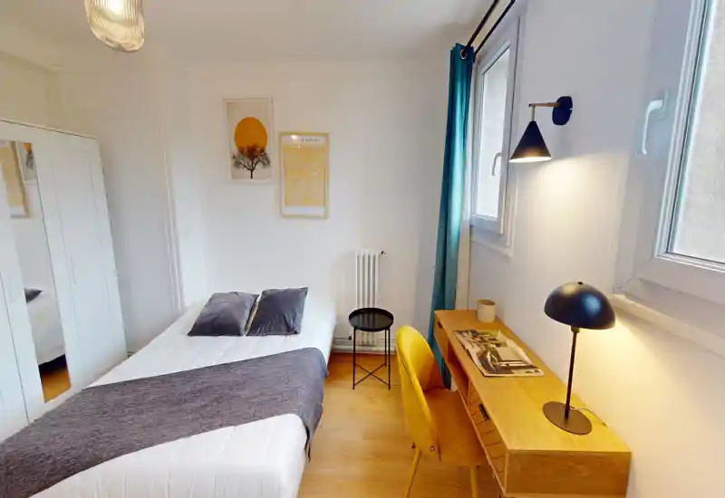 Chambre lumineuse et soignée avec lit double, oreillers moelleux et jeté assorti, bureau en bois avec lampe moderne et fauteuil jaune confortable près de la fenêtre.