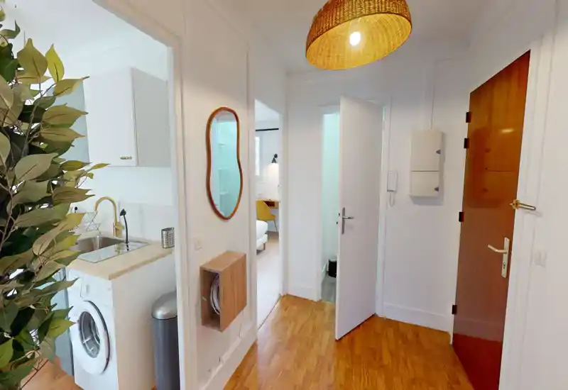 Bright and tidy entry hallway with wooden flooring, rattan pendant lamp and visible apartment door; small kitchenette and laundry area visible to the left.