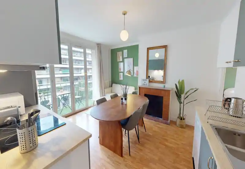 Bright, stylish open-plan living and dining area with a wooden dining table, comfortable chairs, a cosy sofa by a green accent wall, and large glass doors opening to a balcony — ideal for social meals and relaxation.
