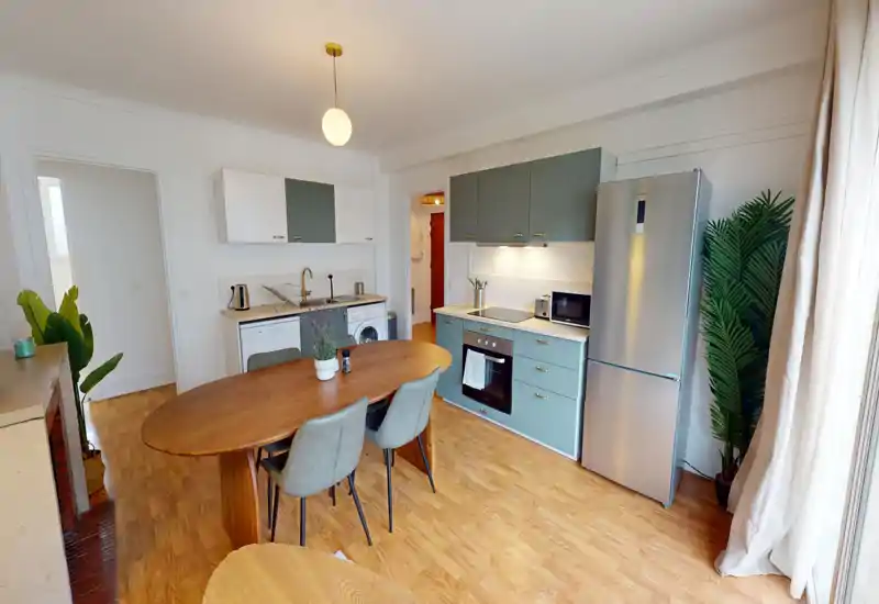 Bright, modern open kitchen with muted green cabinets, stainless fridge, built-in oven and a wooden dining table — ideal for relaxed meals and social cooking.