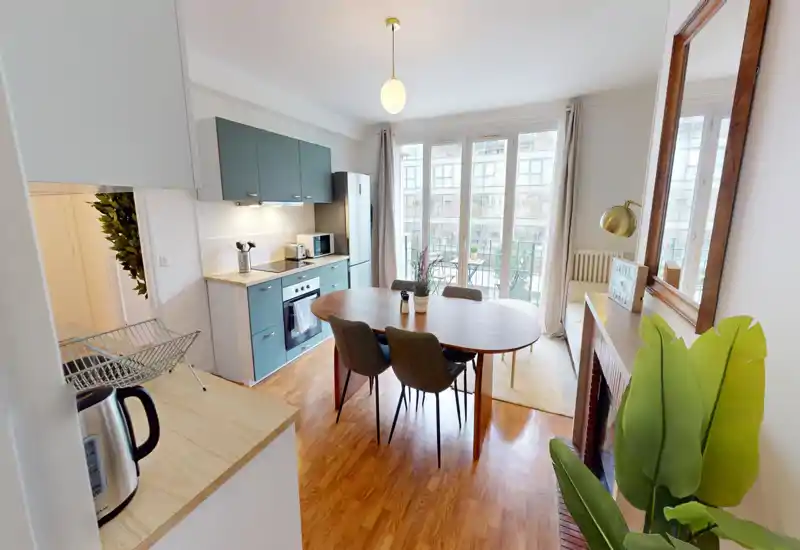 Bright, modern living area with an integrated kitchenette and a central dining table. Large windows and warm wood floors make the space inviting and airy.