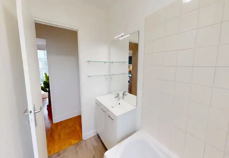 Bright, tidy bathroom with a white vanity, mirror and tiled wall next to a bathtub. Clean modern fixtures and good natural light make this bathroom inviting.
