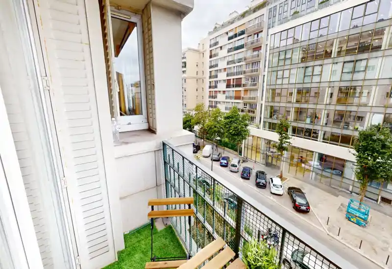 Small furnished balcony overlooking a quiet city street, with folding wooden chairs and a metal railing — ideal for morning coffee.