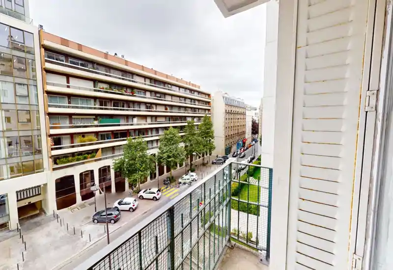 Sunny-looking balcony view over a tree-lined city street with mid-rise residential buildings and parked cars — ideal for apartment listings highlighting an outdoor terrace.