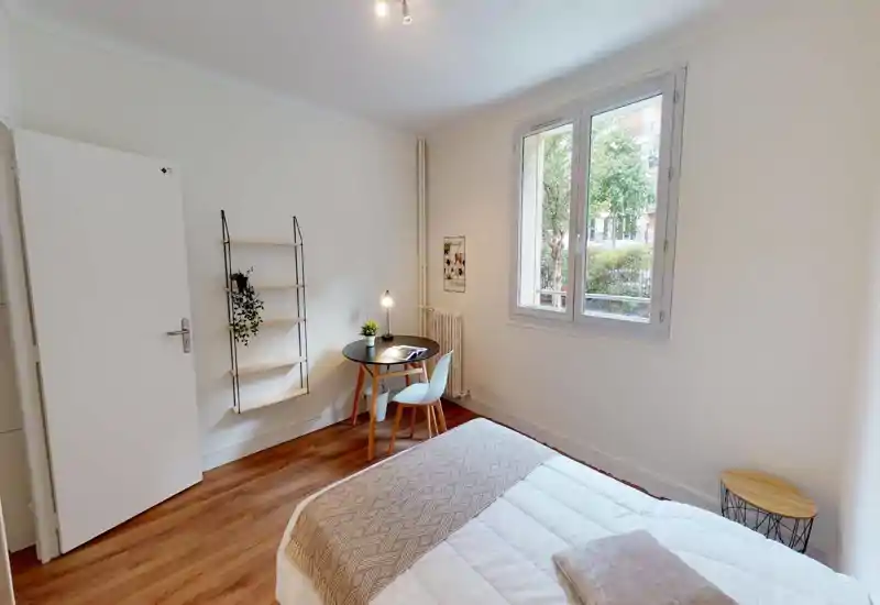 Chambre lumineuse et minimaliste avec un lit au premier plan, une petite table ronde et une chaise près de la fenêtre, et un sol en bois chaleureux. La lumière naturelle et la décoration sobre donnent une atmosphère calme et accueillante.