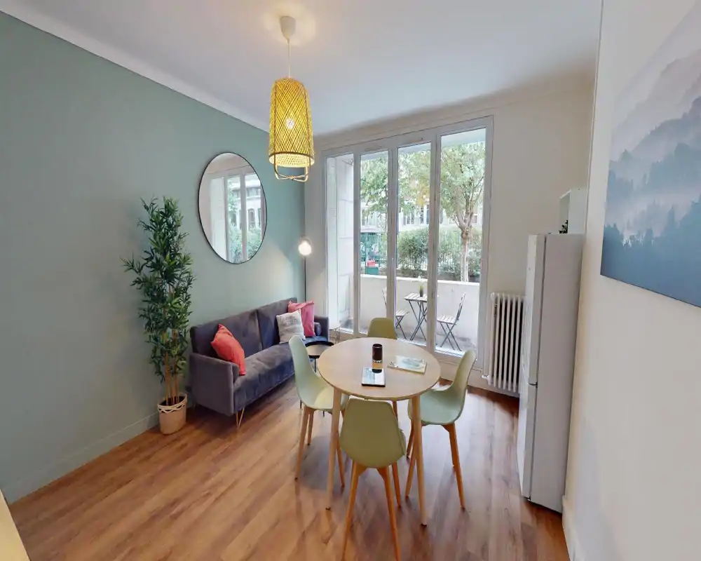 Espace de vie lumineux avec assises, une table basse et des plantes ; de grandes fenêtres laissent entrer la lumière malgré les artefacts de l'image.