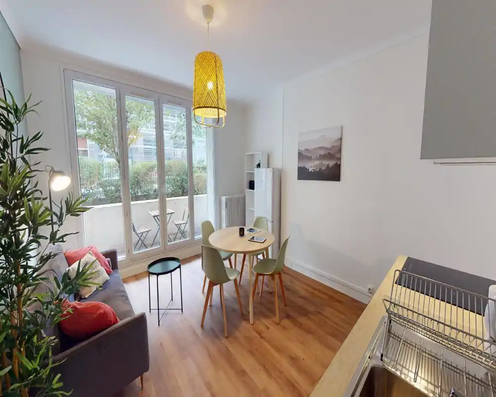 Pièce à vivre moderne et lumineuse avec canapé, table ronde et grandes baies vitrées donnant sur une petite terrasse. Espace bien mis en scène, aéré et idéal pour une annonce locative.