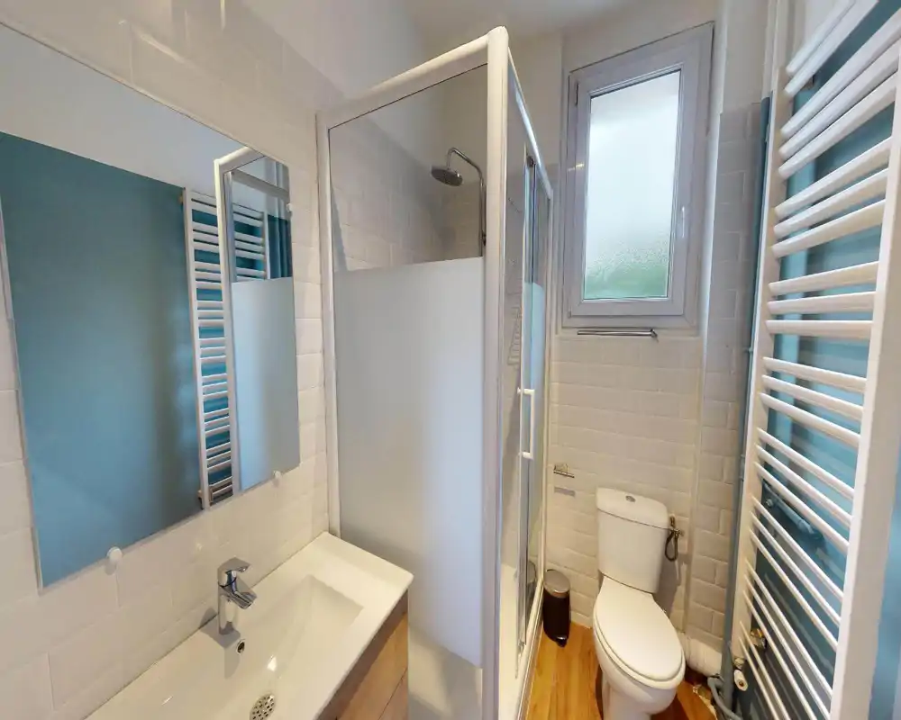 Salle de bain moderne et compacte avec douche d'angle, miroir et lavabo, WC et radiateur sèche-serviettes ; carrelage clair et fenêtre dépoli pour lumière naturelle et intimité.