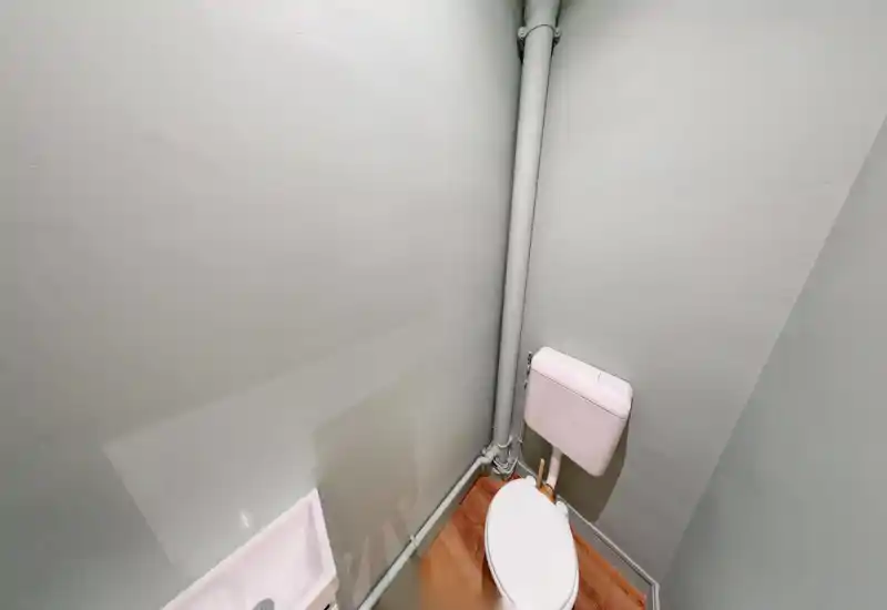 Compact bathroom corner with a wall-mounted cistern and toilet bowl on wood-effect flooring; simple, clean and well-lit.