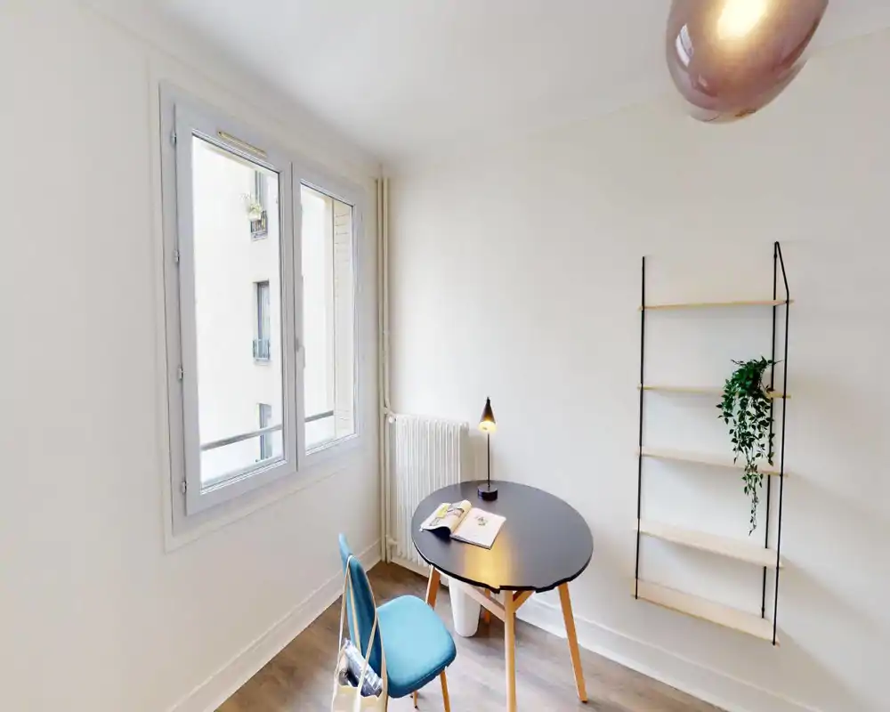 Coin séjour/bureau lumineux et minimaliste avec une table ronde, une chaise et des étagères murales ; une grande fenêtre apporte beaucoup de lumière naturelle.