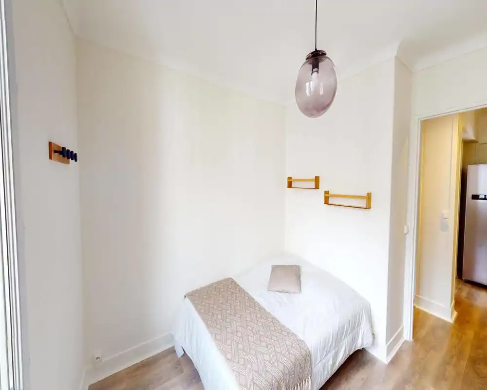 Camera da letto luminosa e minimale con letto matrimoniale ben rifatto, tessili neutri e un elegante lampadario a sospensione. Mensole in legno e pavimento chiaro creano un'atmosfera tranquilla e ariosa, perfetta per affitti brevi.