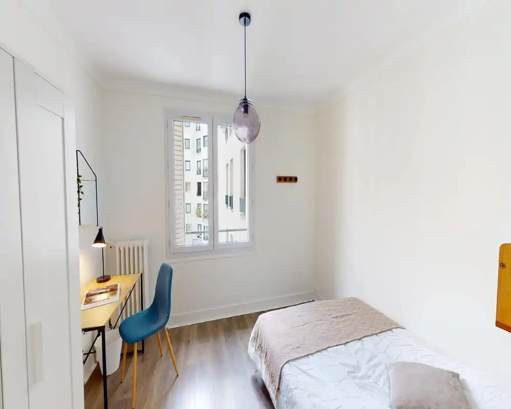 Camera da letto luminosa e minimal con letto singolo in lenzuola neutre, coperta e cuscino, scrivania e sedia vicino alla finestra per un piccolo spazio di lavoro e un moderno lampadario a sospensione.