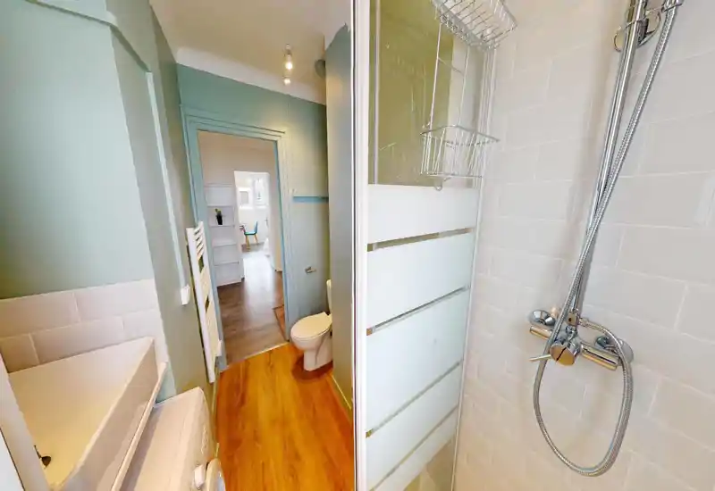 Compact, modern bathroom with a chrome shower head, frosted glass shower door, toilet and sink area; clean white tiles and warm wood-effect flooring give a fresh, tidy feel.