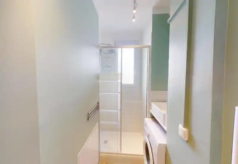 Compact, well-lit bathroom with a glass-enclosed shower, tiled walls and a stacked washing machine beneath a small sink area—functional and tidy.