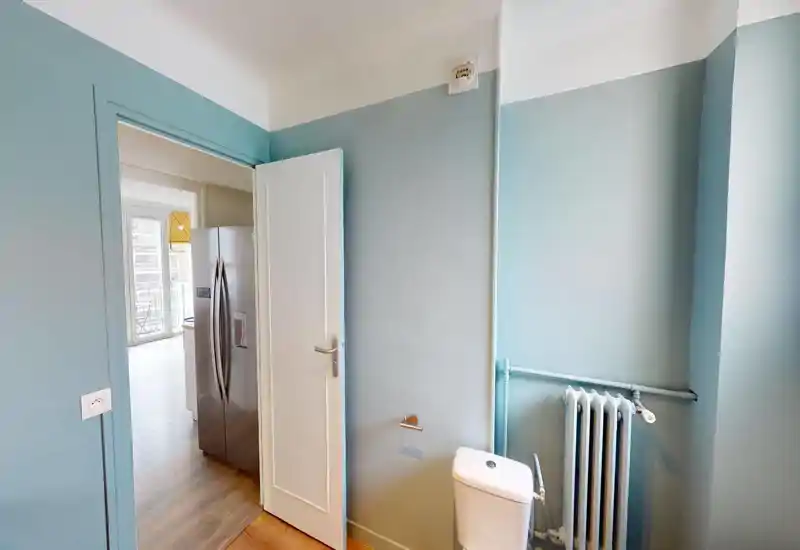 Small bathroom corner with a visible toilet and radiator; soft natural light from an adjacent room highlights the blue-gray painted walls.