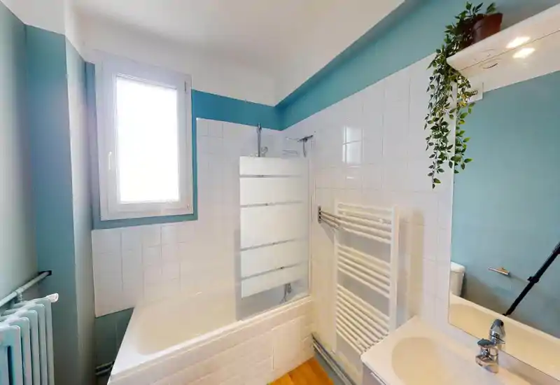 Bright, clean bathroom with a bathtub and overhead shower, white tiled walls and a modern sink. The room features a heated towel rack and a frosted glass shower screen.