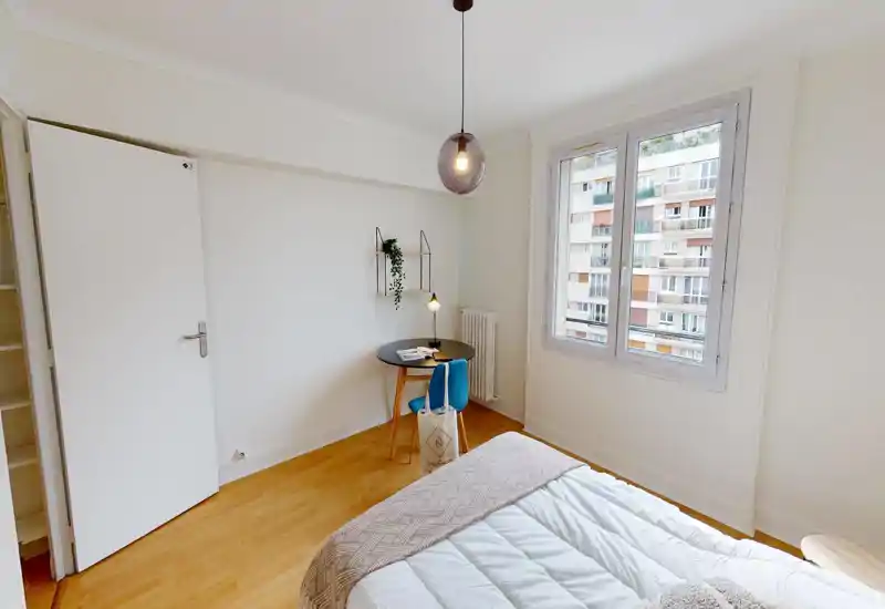 Camera da letto luminosa e minimale con letto ben rifatto in primo piano, piccolo angolo studio vicino alla finestra e pavimento in legno chiaro. Ampia finestra che garantisce molta luce naturale.