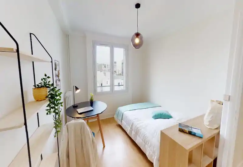Chambre lumineuse et minimaliste avec un lit simple, un petit bureau près de la fenêtre et des éléments décoratifs — idéale pour un étudiant ou une personne seule.