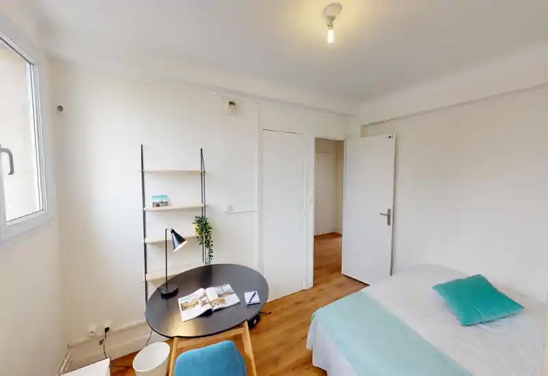 Chambre lumineuse et sobre avec un lit double en linge blanc et un coussin turquoise. Petite table ronde et étagères près de la fenêtre offrant un coin bureau accueillant et lumineux.