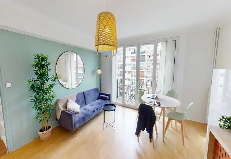 Séjour lumineux et moderne avec un canapé en velours, un miroir rond et une table à manger près de grandes fenêtres donnant sur un petit balcon.