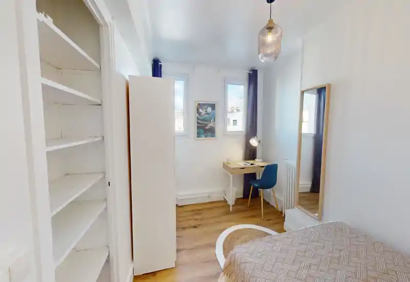 Bright, cozy bedroom with a patterned bedspread, a small desk by two windows and a full-length mirror — ideal for a single occupant or student.