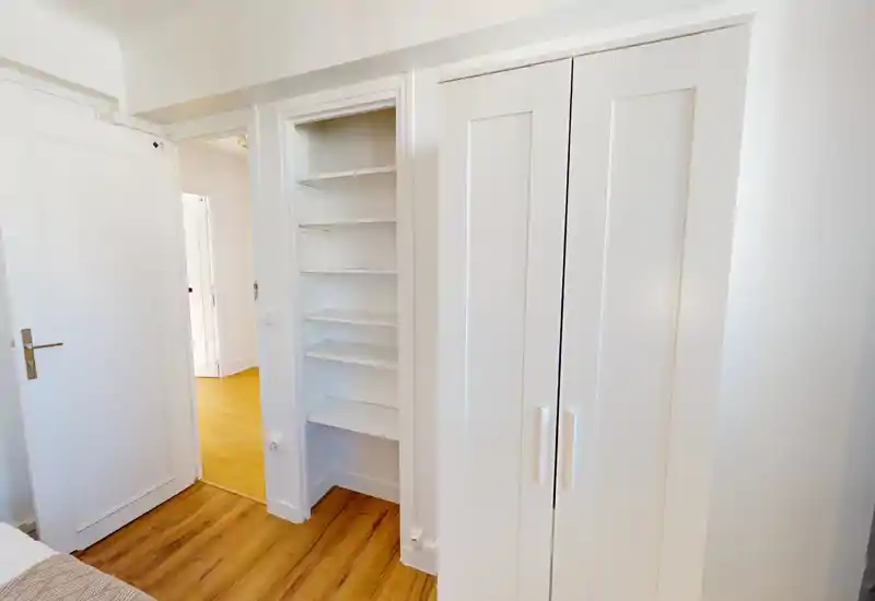 Compact bedroom corner with white built-in shelving and a white wardrobe; warm wood flooring and bright natural light create a clean, minimal look.