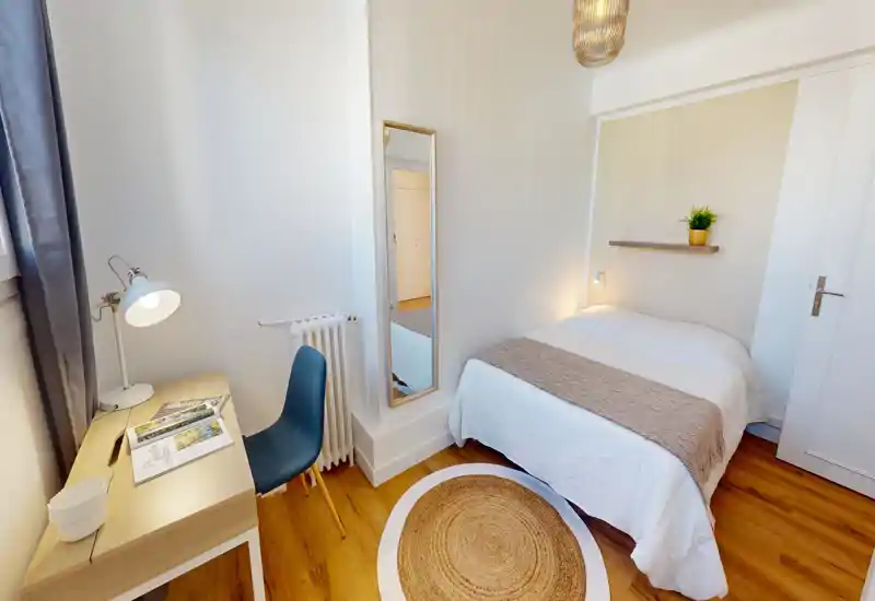 Bright, neatly staged bedroom with a double bed, soft linens, a decorative runner and a small wall shelf with a plant. A desk by the window with a lamp and chair creates a practical work corner; warm wood floors and a round rug add cozy texture.