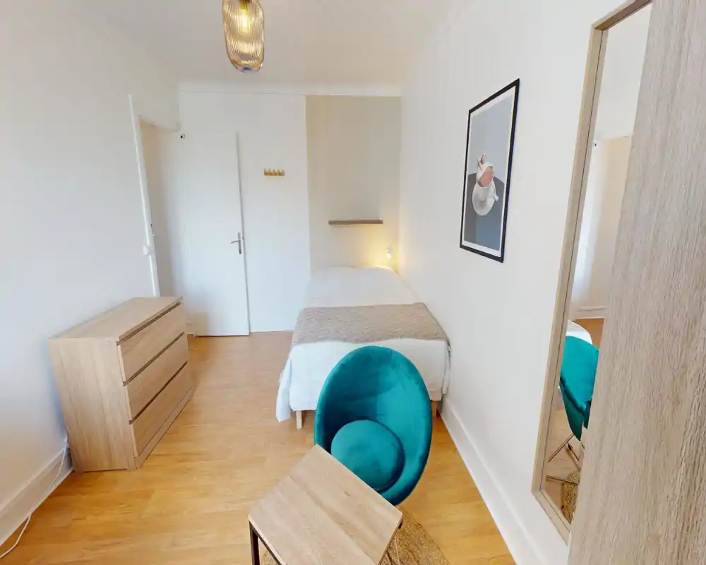 Bright, simply furnished bedroom with a single bed, bedside light and a teal accent chair. Clean wooden flooring and minimal decor create a calm, modern atmosphere ideal for short stays.