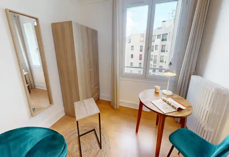 Bright, compact living area with a round wooden table, teal upholstered chairs, a full-height mirror and a wardrobe; large window provides good natural light and warm wooden flooring.