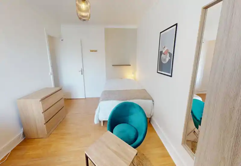 Bright, simply furnished bedroom with a single bed, bedside light and a teal accent chair. Clean wooden flooring and minimal decor create a calm, modern atmosphere ideal for short stays.