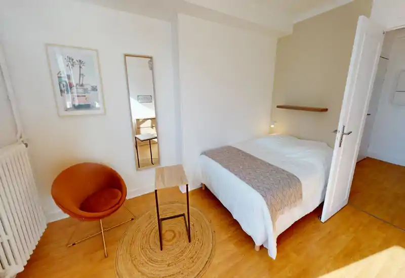 Sunny, well-staged bedroom with a double bed dressed in neutral linens, a small side shelf and a cozy accent chair — ideal for a relaxing stay.