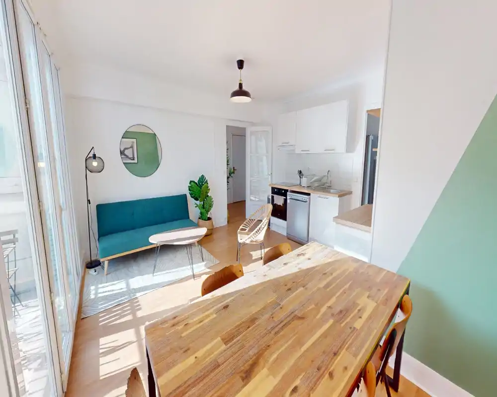 Bright, modern living area with a teal sofa, natural wood dining table and an open kitchenette. Large windows flood the space with light, creating an airy, welcoming atmosphere.