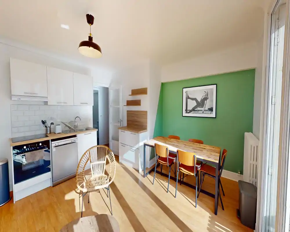 Bright, modern open-plan living area with a compact kitchen and a wooden dining table. Sunlight fills the space, highlighted by a green accent wall and warm wood flooring.
