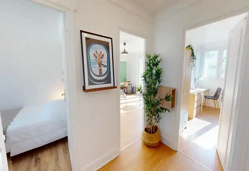 Bright, modern bedroom visible through a doorway: clean white bedding, warm wooden floor and soft natural light create an inviting sleeping area within a well-styled apartment.