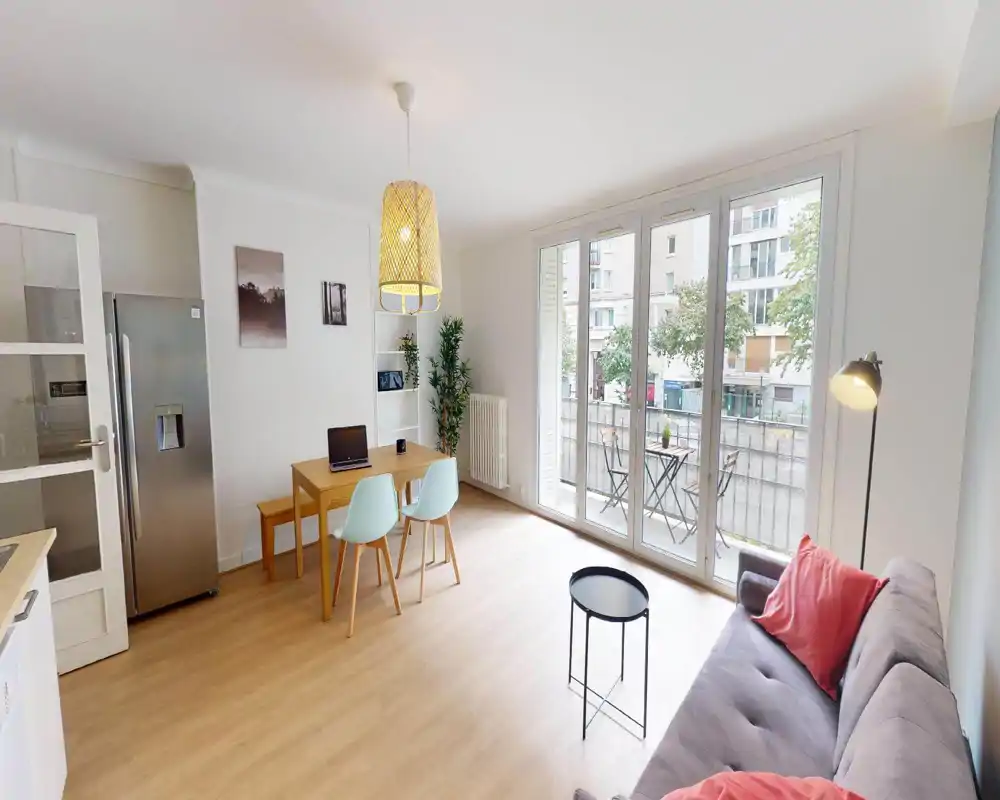 Bright living area with large windows, a sofa and dining chairs; the room shows light wood floors and pendant lighting, offering a modern, airy atmosphere.
