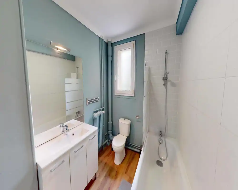 Clean, modern bathroom with bathtub and shower head, toilet and a double-sink vanity; light blue walls and wood-effect flooring create a fresh, airy feel.