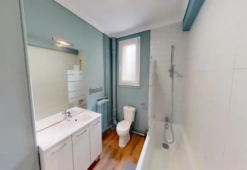 Clean, modern bathroom with bathtub and shower head, toilet and a double-sink vanity; light blue walls and wood-effect flooring create a fresh, airy feel.