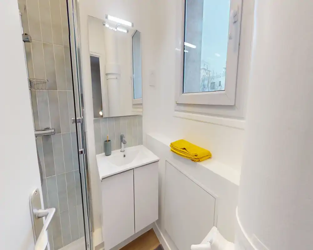 Baño compacto y moderno con mueble lavabo, espejo y ducha en esquina. Acabados blancos y una pequeña ventana que aporta luz natural; una toalla amarilla plegada añade un toque de color.