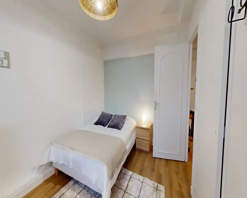 Dormitorio pequeño bien presentado con cama individual, mesita de noche y textiles suaves. Tonos neutros y suelos de madera que crean una atmósfera cálida y acogedora.
