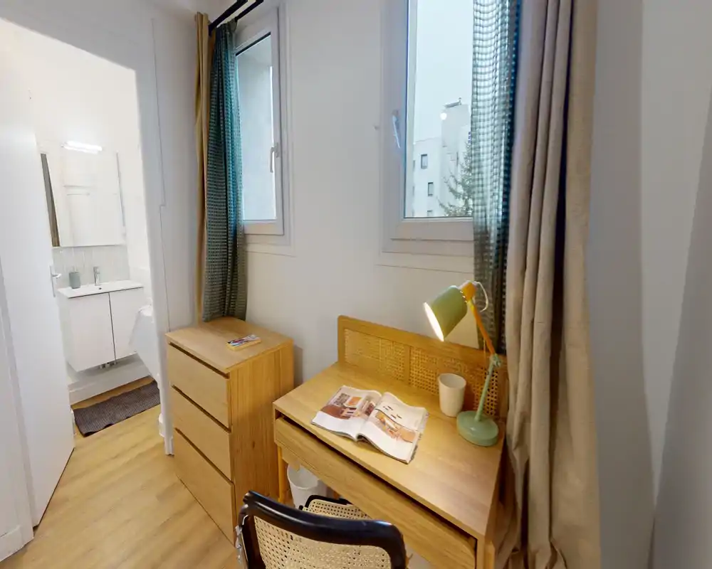 Rincón de dormitorio convertido en zona de estudio: escritorio de madera con lámpara y silla, cómoda de tres cajones a juego y ventanas que proporcionan luz natural. Tonos cálidos de madera y un estilo ordenado crean un espacio compacto y funcional, ideal para una persona o estudiante.