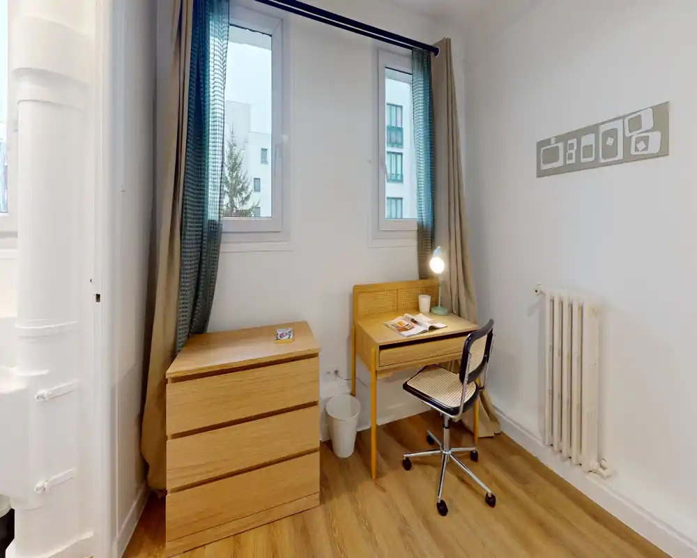 Rincón de habitación luminoso con escritorio de madera, cómoda y silla de oficina con ruedas bajo dos ventanas — ideal como pequeño estudio o espacio junto a la cama.