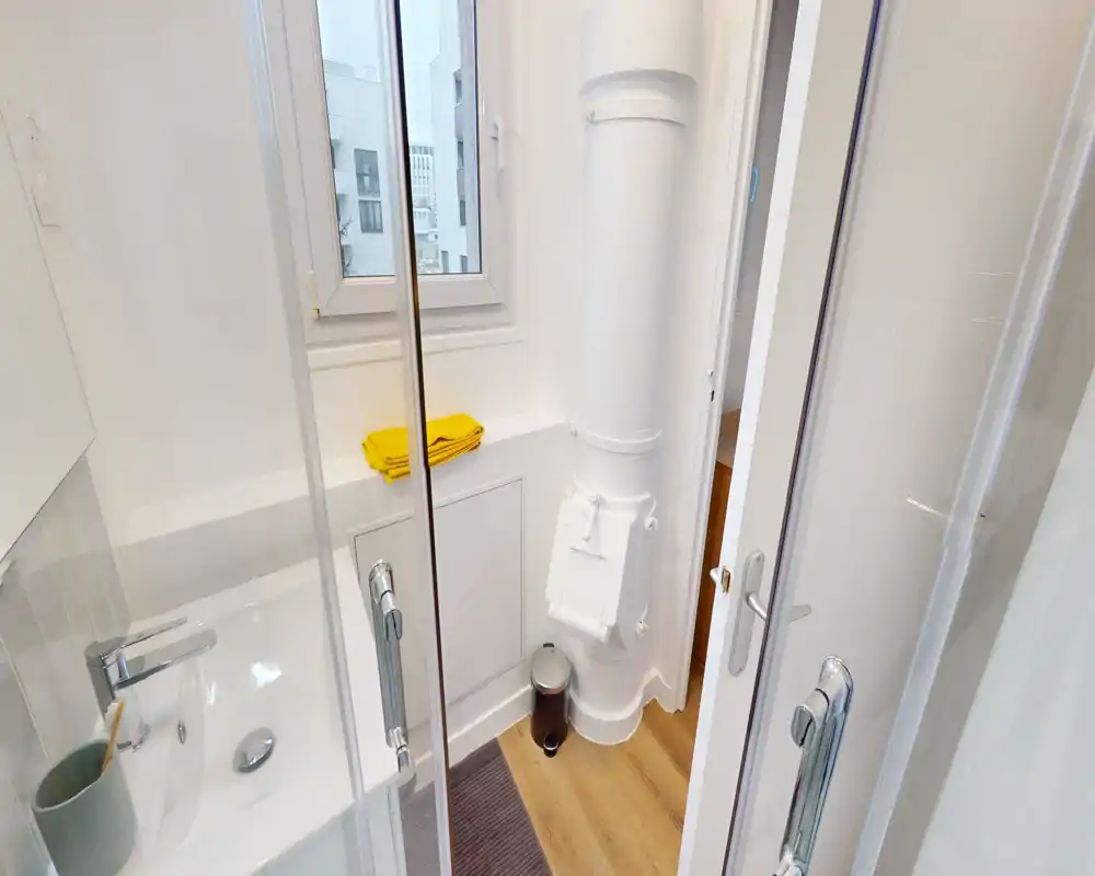 Baño compacto y moderno con azulejos blancos y lavabo junto a la puerta de ducha de vidrio; suelo de madera clara y un pequeño cubo de basura ofrecen contraste.