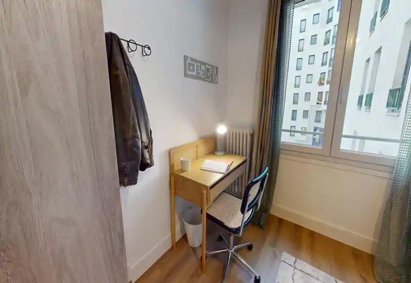 Angolo luminoso con una scrivania compatta in legno e una sedia accanto a una finestra grande; ideale come angolo studio in camera. Pavimento in legno, lampada semplice e giacca appesa che suggeriscono un ambiente abitato ma ordinato.