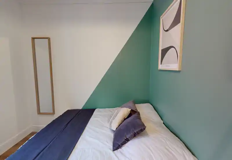 Camera da letto compatta e ben arredata con un letto matrimoniale ordinato contro una parete a due colori e cuscini decorativi — luminosa, moderna e accogliente.