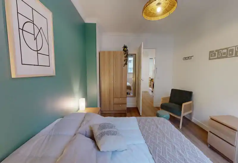 Bright, modern bedroom with a comfortable double bed, decorative cushions and a green accent wall. Natural and artificial lighting highlight wooden furniture including a wardrobe, bedside table and armchair.