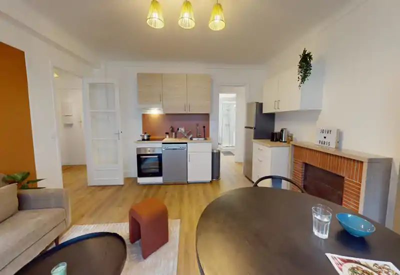 Open-plan living area with a compact fitted kitchen. The space features a sofa, dining table, and modern appliances with warm wood flooring and soft, inviting lighting.
