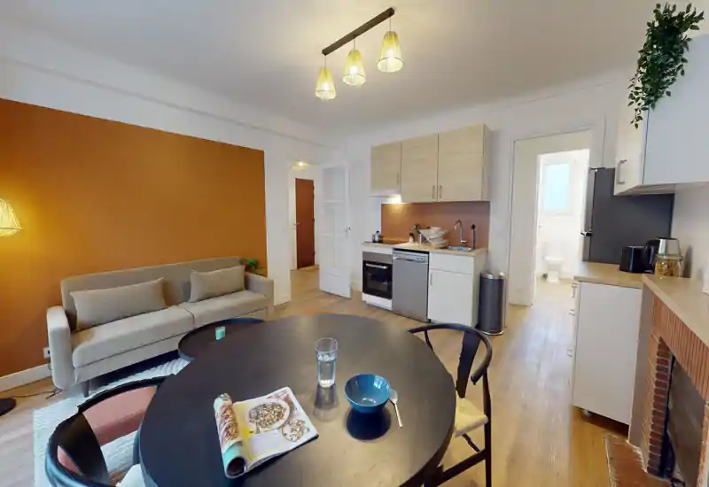 Bright, well-staged open-plan living area with a comfortable sofa, round dining table and a compact kitchenette — warm accent wall and tasteful lighting create a cozy atmosphere.