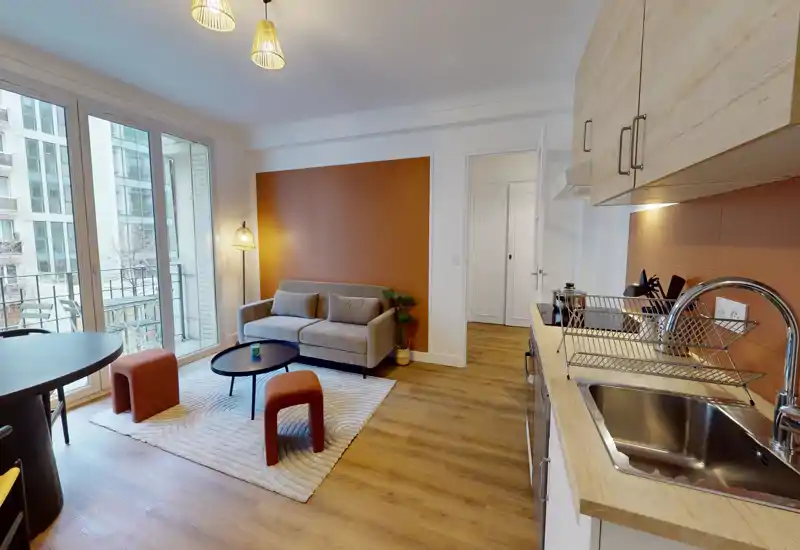 Bright, modern living area with a compact sofa, round coffee table, rug and large windows that open onto a balcony; integrated kitchenette visible to the right, ideal for a city studio.