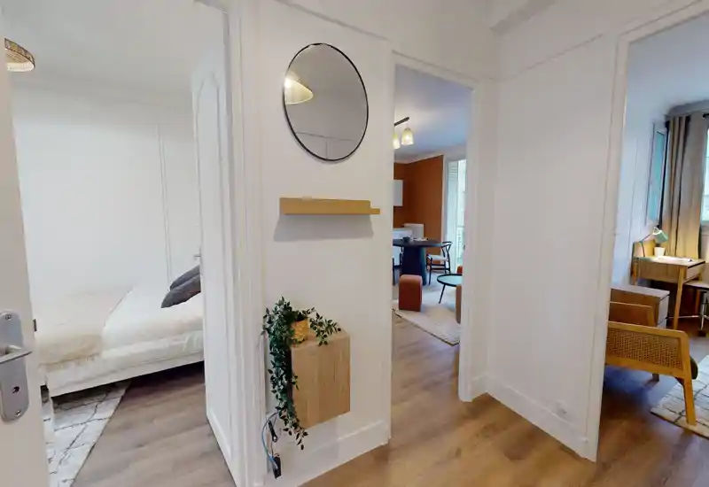 Bright, modern bedroom visible through an open doorway with a white bed and neutral linens; the photo focuses on the hallway and shows adjacent living spaces and tasteful decorative touches.