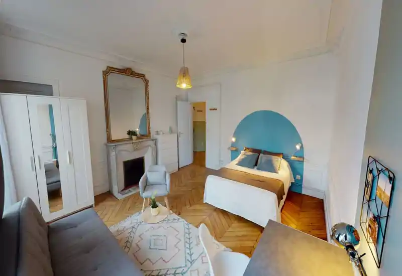 Camera da letto luminosa e raffinata con comodo letto matrimoniale davanti a un semicerchio azzurro come testata, camino classico con specchio decorato, pavimento in parquet e angolo salotto accogliente.